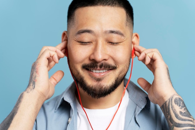 Lächelnder asiatischer Mann mit geschlossenen Augen und roten Kopfhörern, der isoliert auf blauem Grund Musik hört