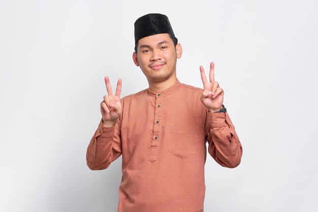 Lächelnder asiatischer moslemischer Mann, der Friedenszeichen mit den Fingern lokalisiert über weißem Hintergrund zeigt