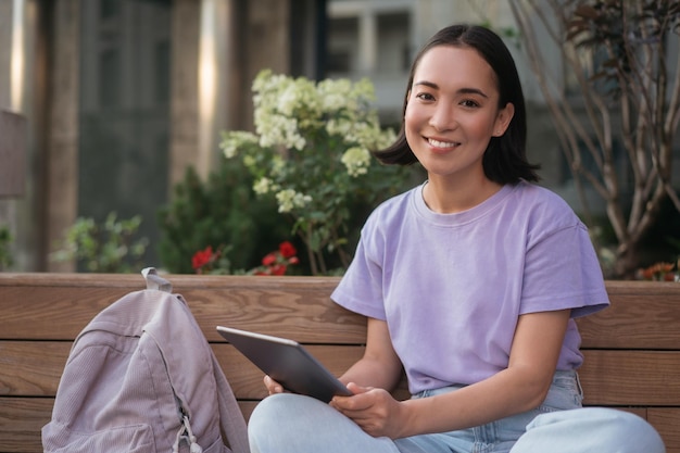 lächelnder asiatischer student, der mit einem digitalen tablet studiert, das auf dem universitätscampus sitzt. Online-Bildung