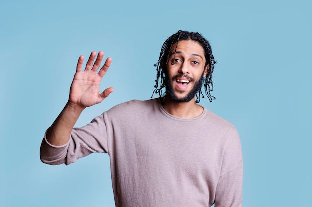 Foto lächelnder attraktiver arabischer mann winkt mit der hand mit fröhlichem gesichtsausdruck. hübsches, freundliches modell mit sorglosen emotionen, das steht und grüßt und mit dem arm begrüßt