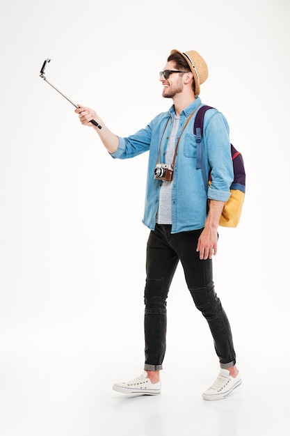 Lächelnder attraktiver junger Tourist mit Rucksack zu Fuß und mit Handy auf Selphie-Stick auf weißem Hintergrund
