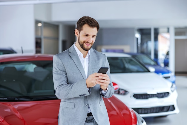 Lächelnder Autoverkäufer, der im Autosalon steht und Telefon benutzt, um auf die Nachricht eines Kunden zu antworten.