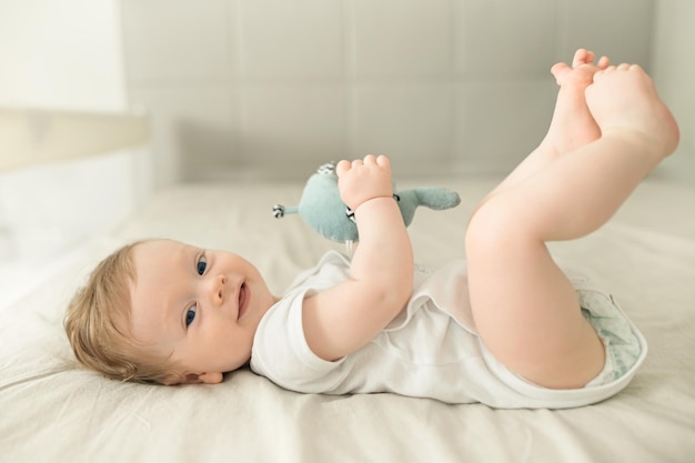 Lächelnder Babyjunge, der mit seinen Beinen und Spielzeug spielt Glückliches blauäugiges Baby, das mit seinen Füßen spielt