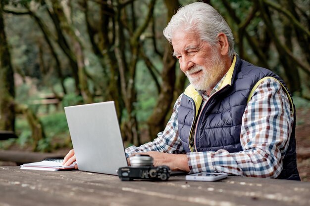 Lächelnder bärtiger älterer Mann, der am Laptop arbeitet, während er draußen im Park an einem Holztisch sitzt