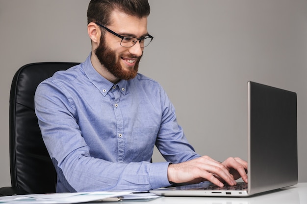 Lächelnder bärtiger, eleganter Mann mit Brille, der etwas in den Laptop schreibt, während er im Büro am Tisch sitzt