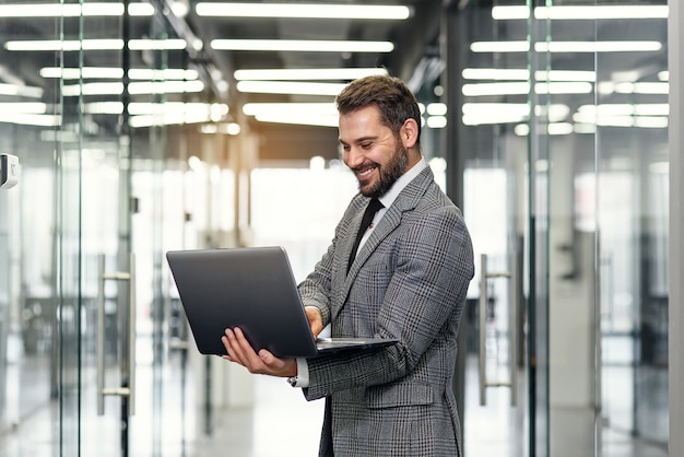 Lächelnder bärtiger Geschäftsmann konzentrierte sich auf Unternehmensdaten, während er im Bürokorridor stand und Laptop verwendete.
