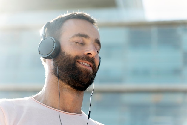 Lächelnder bärtiger Mann, der Musik hört und stylische Kopfhörer trägt, die auf der Straße stehen, Kopierraum