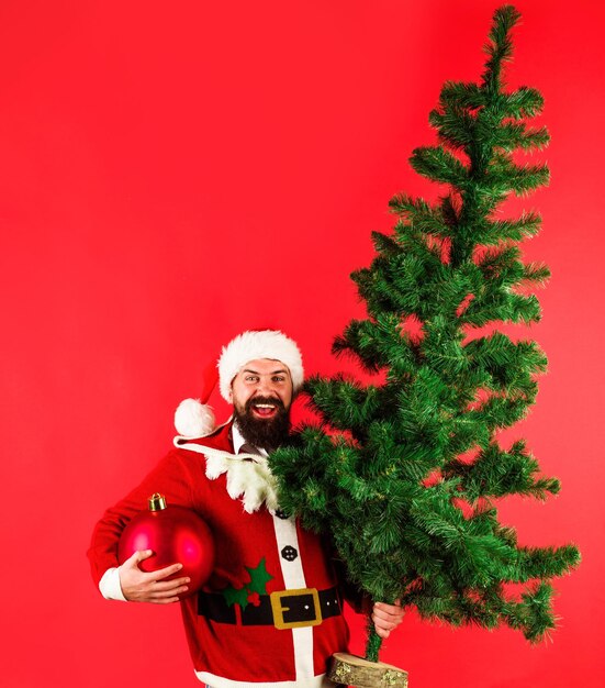 Lächelnder bärtiger mann im weihnachtsmannkostüm mit tannenbaum und großer weihnachtsballwerbung des neuen jahres