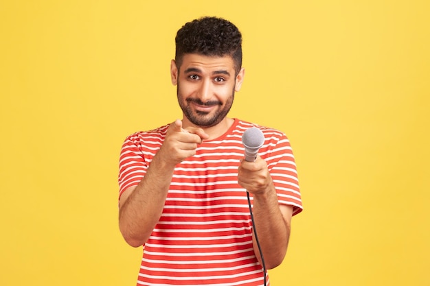 Lächelnder bärtiger Mann in gestreiftem rotem T-Shirt, der ein Mikrofon hält und mit dem Finger auf die Kamera zeigt und Sie auffordert, zu singen oder zu gratulieren Studioaufnahme im Innenbereich isoliert auf gelbem Hintergrund