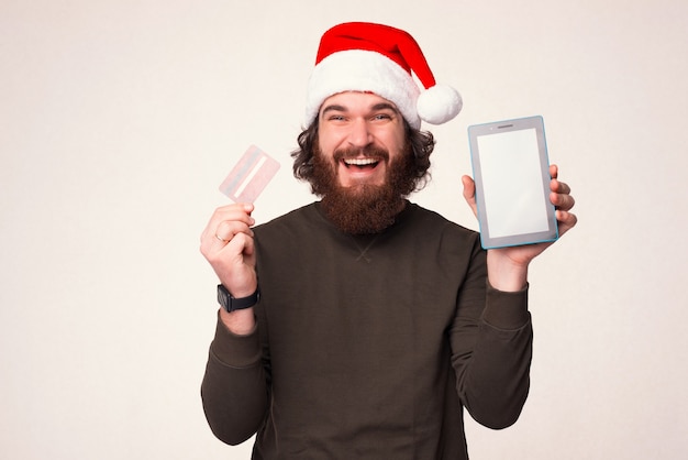 Lächelnder bärtiger Mann mit Weihnachtsmütze hält Bankkarte und sein Tablet.