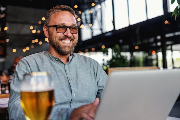 Lächelnder bärtiger Mann mittleren Alters mit Brille, die in seiner Bar sitzt, Laptop benutzt und ein Glas frisches kaltes helles Bier hat.