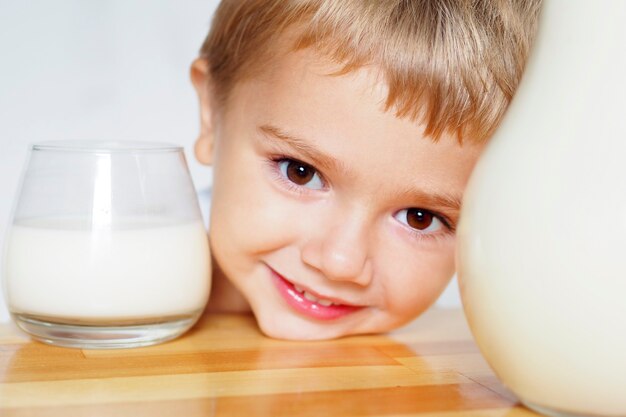 Lächelnder blonder Junge mit braunen Augen trinkt Milch aus einem Glas an einem Holztisch auf weißem Hintergrund.