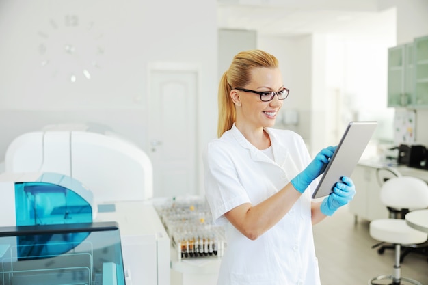 Lächelnder blonder Laborassistent mit Gummihandschuhen, die Tablette halten und Testergebnisse eingeben.