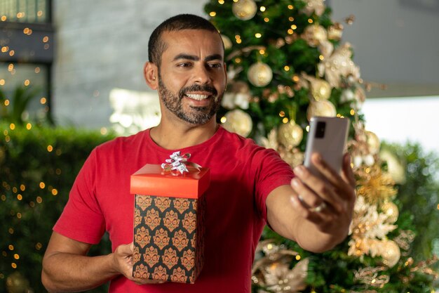 Lächelnder brünette Mann mit roter Bluse und Geschenkbox in den Händen Videoanrufe am Weihnachtstag