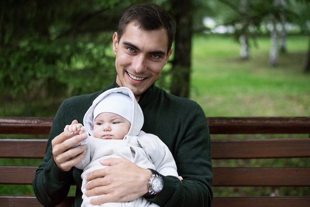 Lächelnder brünetter Vater in reen Jacke sitzt auf Bank mit Baby in seinen Armen im Park, glückliches Vaterschaftskonzept