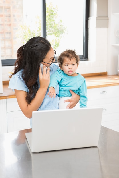 Lächelnder Brunette, der ihr Baby hält und Laptop beim Telefonanruf in der Küche verwendet