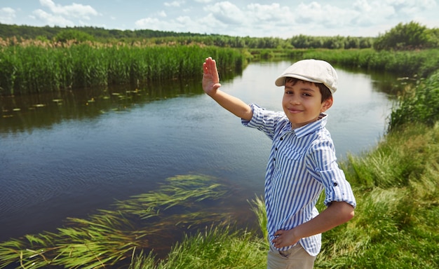 Lächelnder entzückender Junge, der hallo winkt und auf dem Hintergrund des Flussufers steht. Kinderjunge im Sommerurlaub auf dem Land