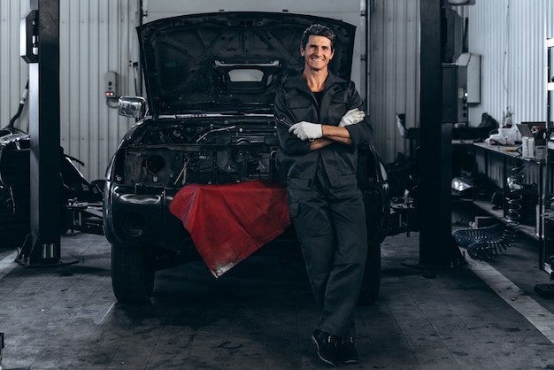 Lächelnder erwachsener Mann in Arbeitsuniform, der in der Nähe des schwarzen offenen Autos in der großen Autogarage posiert. Auto-Check-up-Konzept