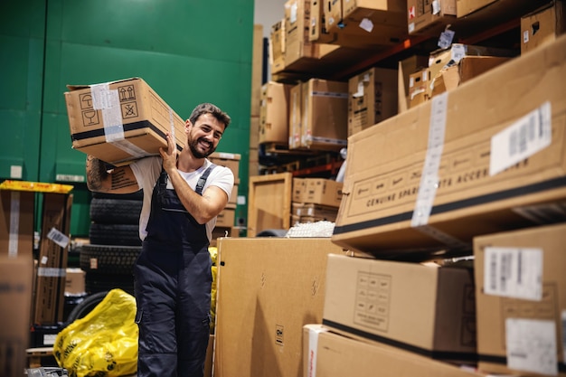 Lächelnder fleißiger tätowierter bärtiger Arbeiter in Overalls, der eine Schachtel auf seiner Schulter trägt und sie für den Export vorbereitet, während er durch das Lager geht.