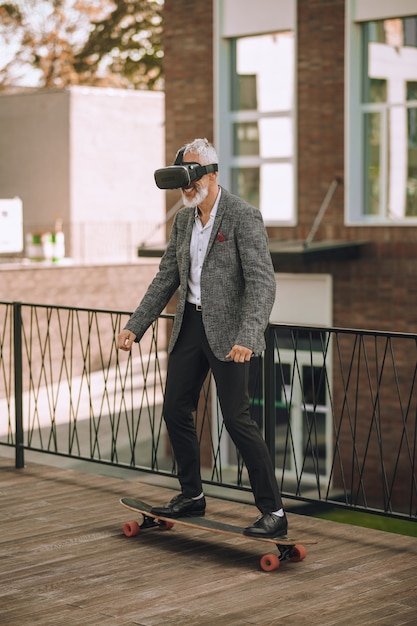 Lächelnder fröhlicher bärtiger grauhaariger Mann im Virtual-Reality-Headset, der mit beiden Füßen auf dem Brett steht