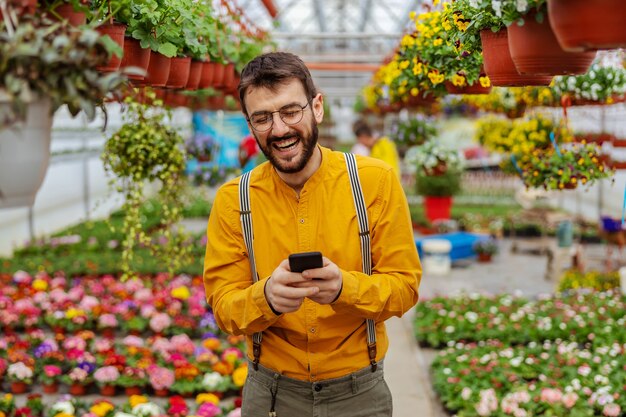 Lächelnder Gärtner, der im Gewächshaus steht und Bestellungen von einem Kunden über Smartphone entgegennimmt.