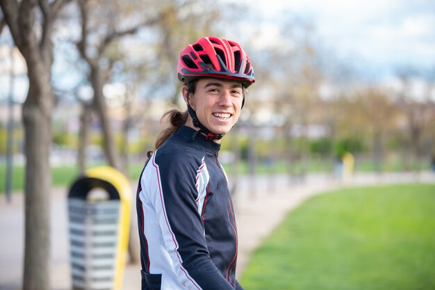 Lächelnder geeigneter männlicher Radfahrer der Junge in der Sportkleidung und in Sturzhelm, die auf Fahrrad im Park sitzen und Kamera betrachten