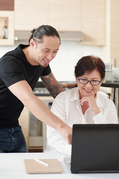 Lächelnder gemischtrassiger Mann, der Mutter erklärt, wie man einen neuen Laptop benutzt und nützliche Informationen online findet