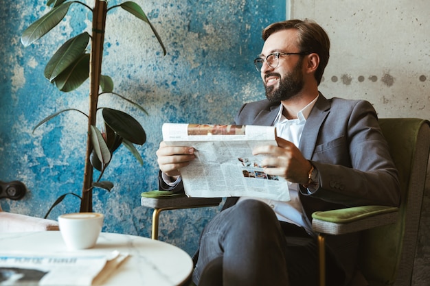 Lächelnder Geschäftsmann, der Anzug liest Zeitung liest, während er im Café sitzt und Kaffee trinkt