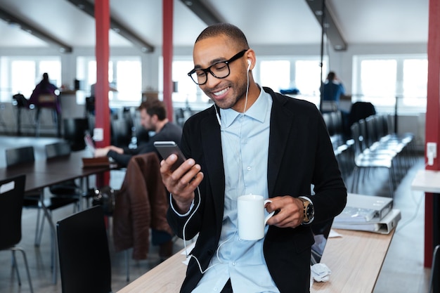 Lächelnder Geschäftsmann, der eine SMS auf dem Smartphone schreibt und eine Tasse im Büro hält