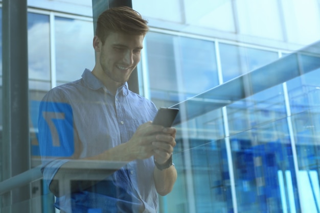 Lächelnder Geschäftsmann, der im Büro steht und Handy benutzt