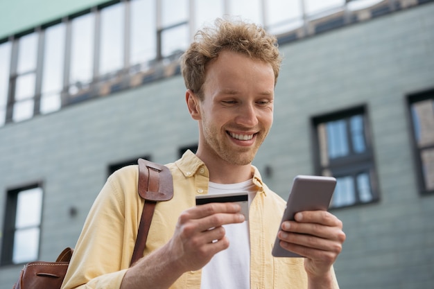 Lächelnder Geschäftsmann, der Kreditkarte mit Smartphone für Online-Shopping auf der Straße hält
