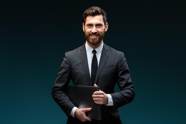 Lächelnder Geschäftsmann, der Laptop in den Händen hält, während er mit Freude in die Kamera schaut, lächelt isoliert auf dem blauen Studiohintergrund