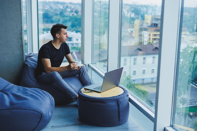 Lächelnder Geschäftsmann, der nach etwas sucht und auf einem Laptop tippt und frischen schwarzen Kaffee trinkt Das Konzept einer modernen erfolgreichen Person Junge fokussierte Kerl mit Gläsern in einem Großraumbüro