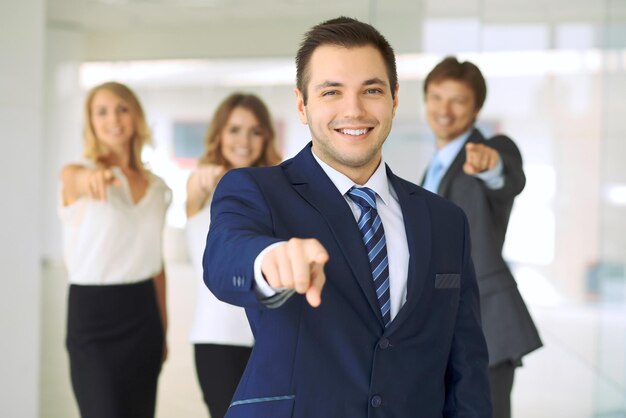 Lächelnder Geschäftsmann im Büro mit Kollegen im Hintergrund, der mit dem Finger in die Kamera zeigt