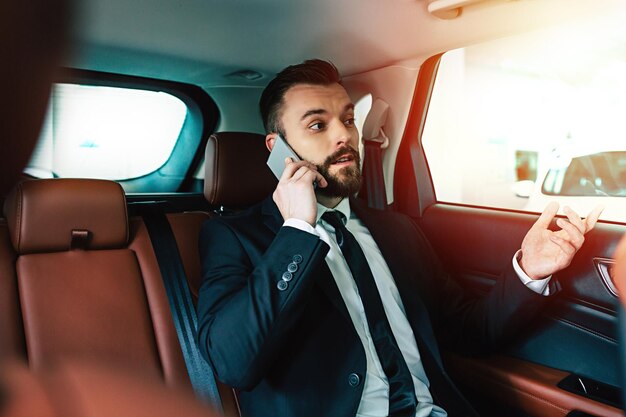 Lächelnder Geschäftsmann im vollen Anzug, der am Telefon spricht, während er auf dem Rücksitz eines Taxiautos sitzt