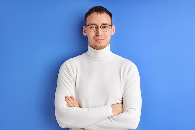 Lächelnder Geschäftsmann in Brillen, die Kamera mit verschränkten Armen betrachten, tragen weißes Freizeithemd, lokalisiert auf blauem Studiohintergrund