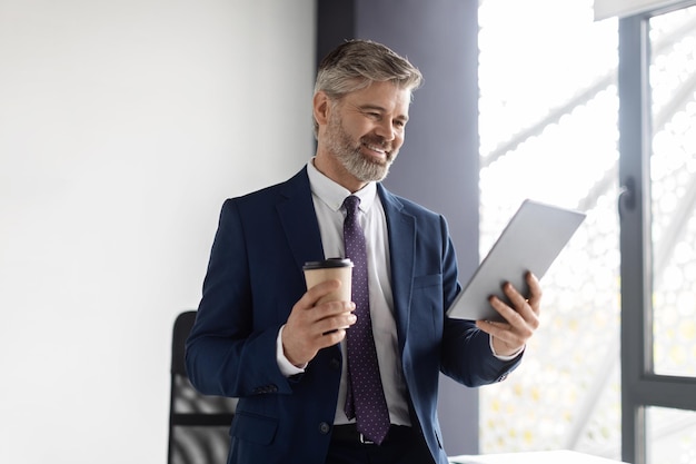 Lächelnder Geschäftsmann mittleren Alters, der Kaffee trinkt und im Büro ein digitales Tablet verwendet