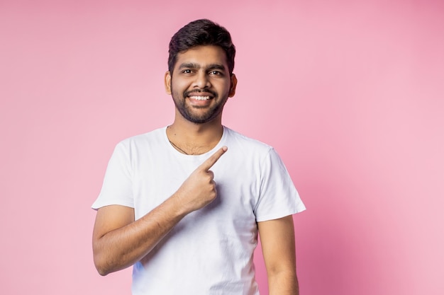 Lächelnder, glücklicher, bärtiger junger Mann, der mit dem Zeigefinger nach oben zeigt, gekleidet in ein weißes, lässiges T-Shirt, das neue Artikel annonciert, einzeln auf rosafarbenem Hintergrund. Werbung, Gestenkonzept.
