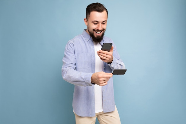 Lächelnder glücklicher gutaussehender Mann, der Alltagskleidung trägt, isoliert auf der Hintergrundwand, die Telefon und Kreditkarte hält und die Zahlung mit Blick auf den Smartphone-Bildschirm macht