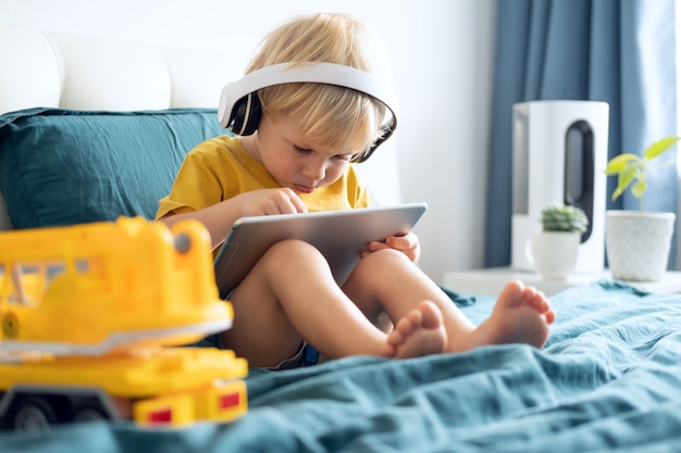 Lächelnder glücklicher kleiner Junge mit Tablet in drahtlosen Kopfhörern im Bett zu Hause.