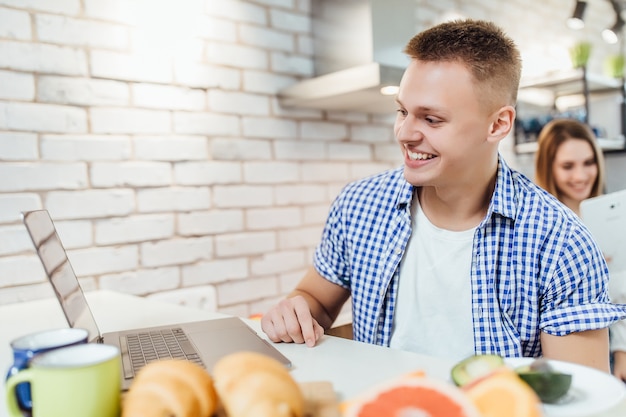 Lächelnder glücklicher Mann, der das Frühstück in der Küche genießt und auf den Laptop-Bildschirm schaut, der Videoanrufe tätigt,