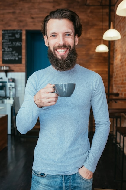 Lächelnder glücklicher Mann, der in der Kaffeestube steht, die Tasse des heißen Getränks hält