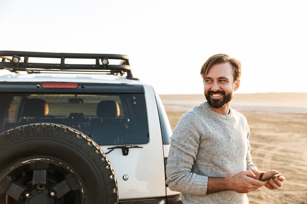 Lächelnder gutaussehender junger Mann mit Handy beim Stehen am Auto am sonnigen Strand