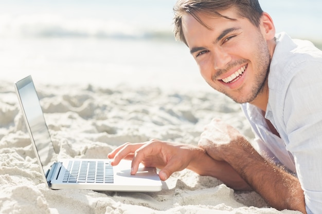 Lächelnder gutaussehender Mann auf dem Strand unter Verwendung seines Laptops