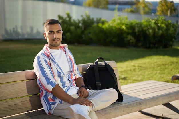 Lächelnder, gutaussehender Mann aus dem Nahen Osten mit Rucksack, der in die Kamera blickt, die draußen auf einer Bank sitzt