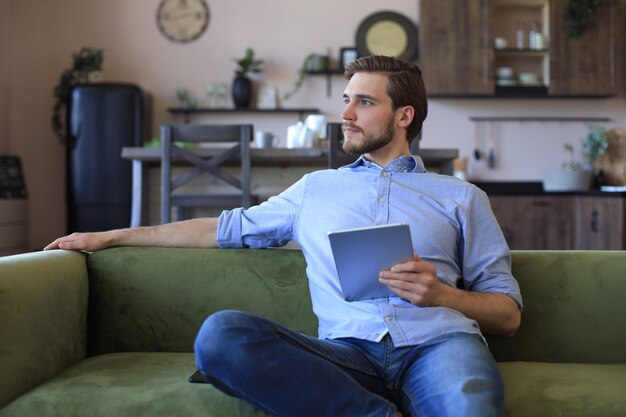 Lächelnder gutaussehender Mann, der zu Hause ein Tablet mit sozialen Netzwerken hält und verwendet, glücklicher Freiberufler, der Online-Shopping macht, auf einem gemütlichen Sofa im Wohnzimmer sitzt.