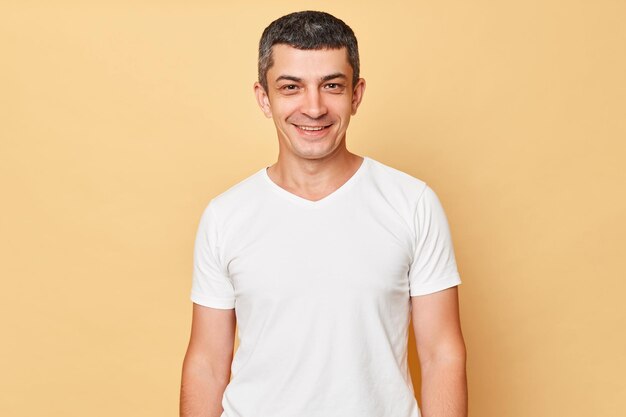 Lächelnder, gutaussehender Mann in weißem, lässigem T-Shirt, der isoliert vor beigem Hintergrund steht und in die Kamera blickt, gut gelaunt ist und positive Gefühle zum Ausdruck bringt