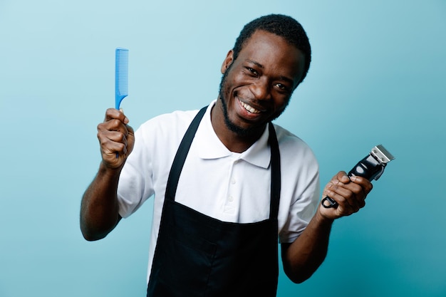 Lächelnder Haltekamm mit Haarschneidemaschine junger afrikanisch-amerikanischer Friseur in Uniform isoliert auf blauem Hintergrund