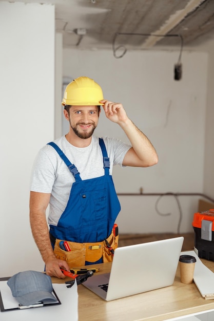 Lächelnder Handwerker in Uniform mit Werkzeuggürtel und Hardhat, der während der Renovierungsarbeiten im Innenbereich in die Kamera schaut