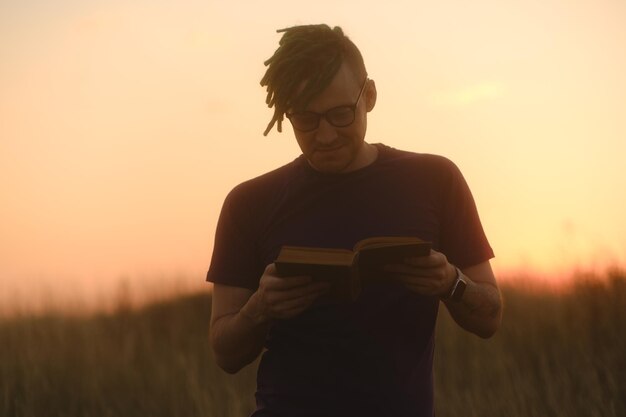 Lächelnder Hipster mit Buch Niedriger Winkel eines jungen Mannes mit Dreadlocks in legerer Kleidung und Brille, der mit einem Buch vor Sonnenuntergang steht